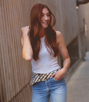 Wearing the Dreamweaver woven leather belt by Bed|Stü, a person with long auburn hair in a white sleeveless top and jeans stands outdoors, smiling with one hand in their pocket—the artisan craftsmanship of the belt perfectly accentuating their bohemian style.