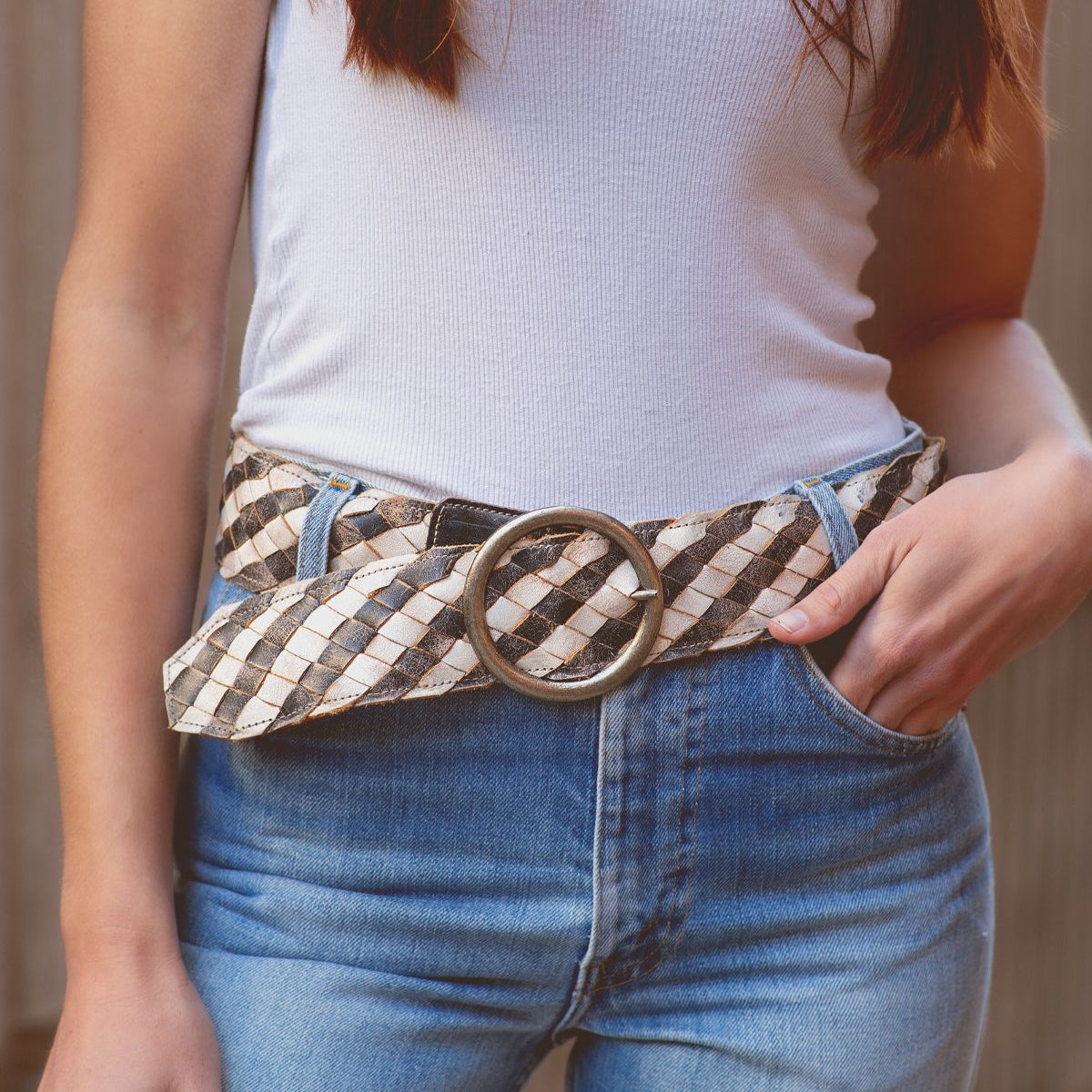 Wearing the Bed|Stü Dreamweaver—a woven leather belt with a large round buckle—paired with a white tank top and blue jeans, this person keeps one hand in their pocket for a perfect touch of bohemian style.