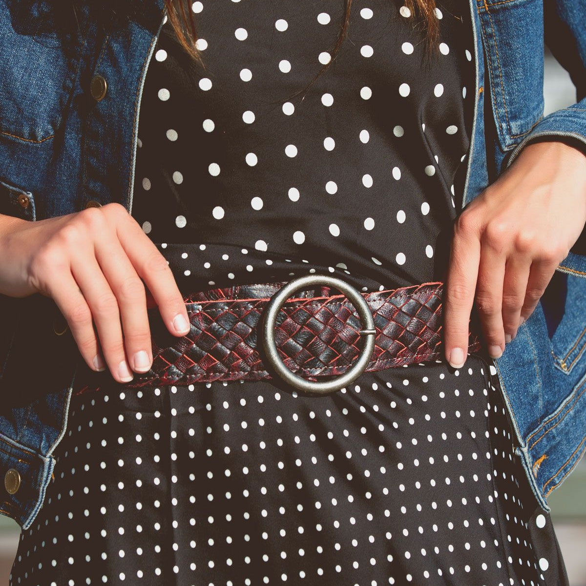 A woman wears a black and white polka dot dress, a denim jacket, and the Bed|Stü Dreamweaver—a wide woven leather belt with a large round buckle that showcases artisan craftsmanship.