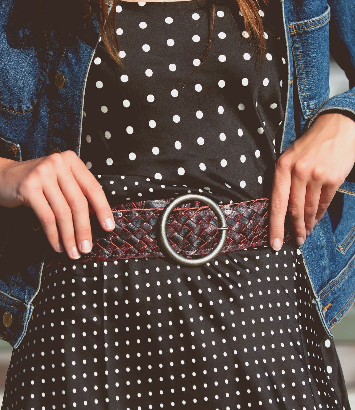 A woman wears a black and white polka dot dress, a denim jacket, and the Bed|Stü Dreamweaver—a wide woven leather belt with a large round buckle that showcases artisan craftsmanship.