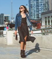 A woman in sunglasses and a denim jacket wears a black polka dot dress, cinched at the waist with the Bed|Stü Dreamweaver woven leather belt, as she walks on a rooftop with city buildings in the background.