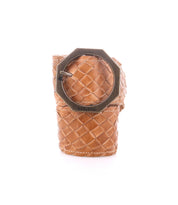 The Dreamweaver napkin ring by Bed|Stü features tan woven leather, reminiscent of a finely crafted belt, and is accented with an octagonal metal buckle engraved with "BED|STÜ." Showcasing artisan craftsmanship and subtle bohemian style, it is displayed on a plain white background.