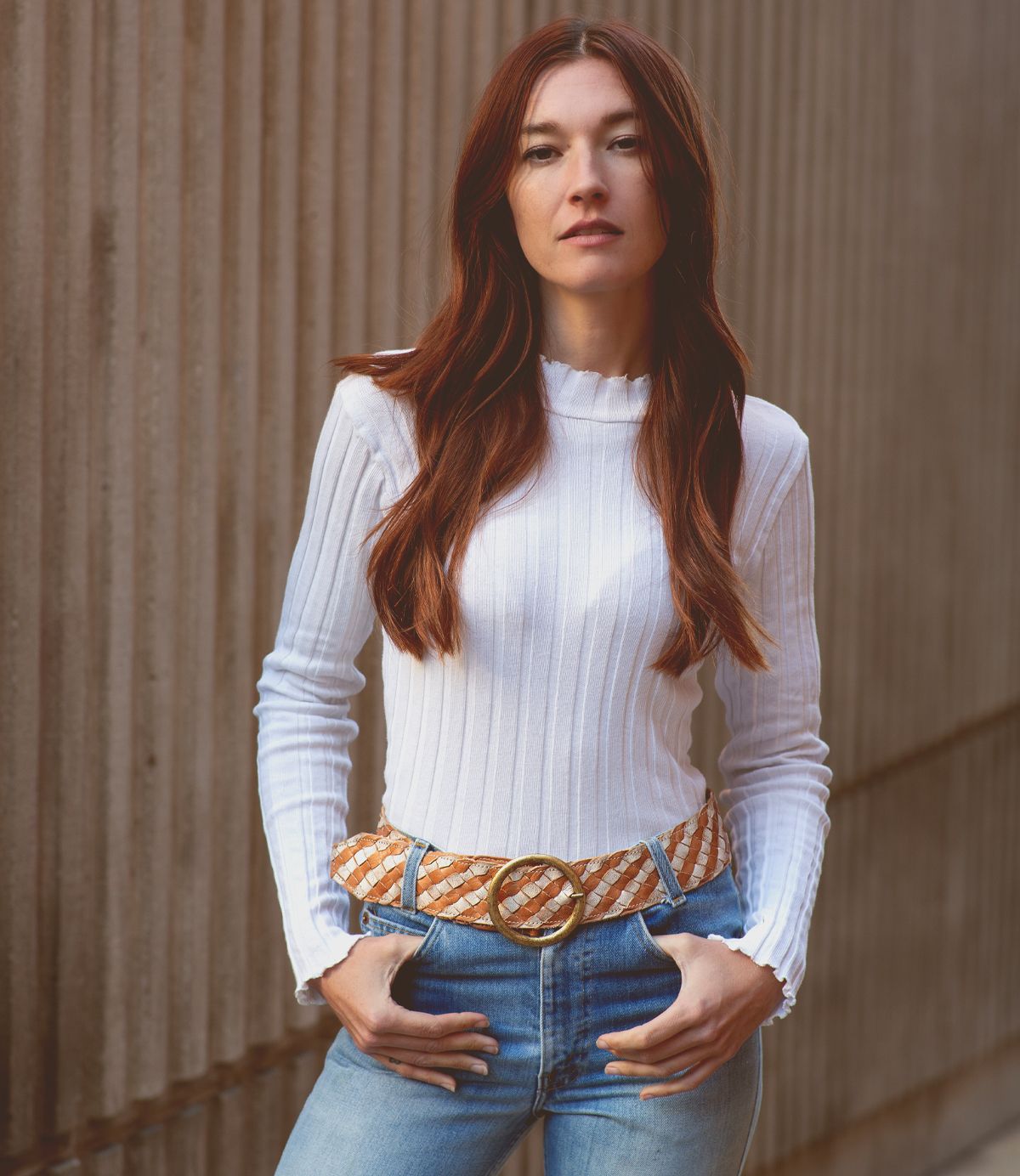 A woman with long brown hair wears a white ribbed top, blue jeans, and the Bed|Stü Dreamweaver woven leather belt, standing outdoors against a textured concrete wall and effortlessly capturing a touch of bohemian style.