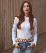 A woman with long brown hair wears a white ribbed top, blue jeans, and the Bed|Stü Dreamweaver woven leather belt, standing outdoors against a textured concrete wall and effortlessly capturing a touch of bohemian style.