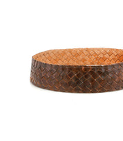 The Dreamweaver basket by Bed|Stü is a round, shallow piece featuring a woven pattern of brown and tan strips that highlights artisan craftsmanship and bohemian style, beautifully set against a plain white background.
