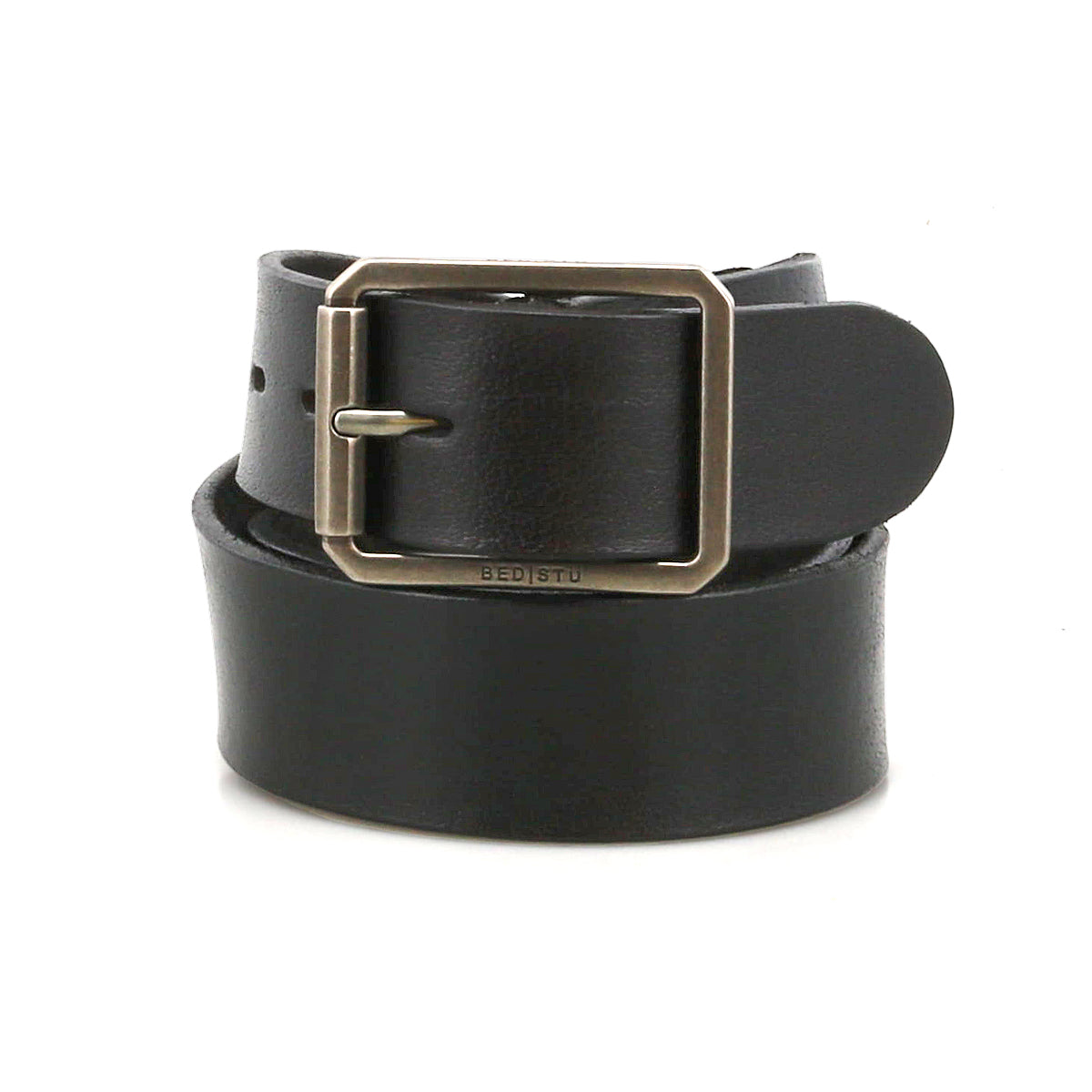 The Drifter by Bed|Stü is a black leather belt featuring a rectangular metal buckle, coiled in a circular shape against a plain white background. Its textured leather strap and minimal design make this Bed|Stü accessory truly timeless.