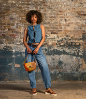 A person stands in front of a textured brick wall, wearing a sleeveless denim top, matching jeans, and sandals, carrying the Elect shoulder bag by Bed|Stü—a small brown accessory that adds vintage charm perfect for everyday use.
