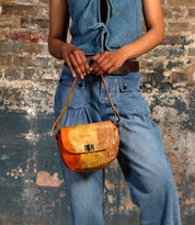 A person in a sleeveless denim top and jeans holds the Bed|Stü Elect, a small tan leather crossbody bag with vintage charm, standing in front of a textured brick wall.