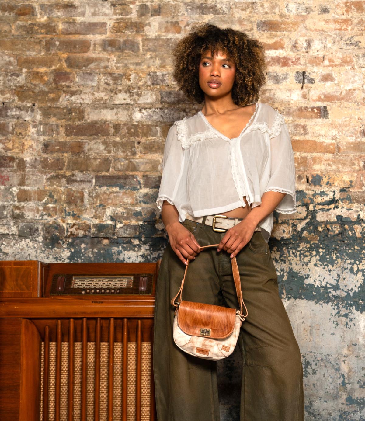 A woman in a white blouse and olive pants leans against a vintage radio in front of a worn brick wall, holding the Bed|Stü Elect shoulder bag—a tan and white accessory that radiates vintage charm, perfect for everyday use.