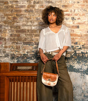A woman in a white blouse and olive pants leans against a vintage radio in front of a worn brick wall, holding the Bed|Stü Elect shoulder bag—a tan and white accessory that radiates vintage charm, perfect for everyday use.