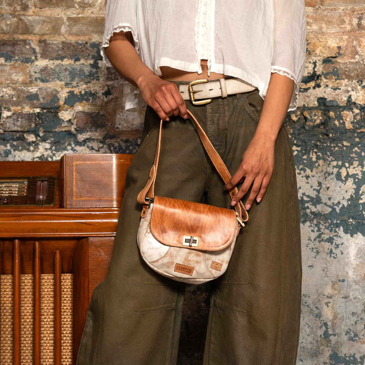 A person in a white blouse and olive pants holds the Bed|Stü Elect, a small brown and white shoulder bag, standing in front of a weathered brick wall and vintage wooden radio, capturing an effortless sense of vintage charm.