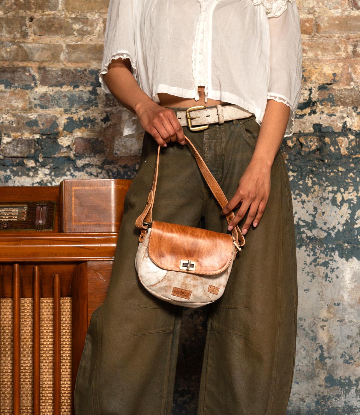A person in a white blouse and olive pants holds the Bed|Stü Elect, a small brown and white shoulder bag, standing in front of a weathered brick wall and vintage wooden radio, capturing an effortless sense of vintage charm.
