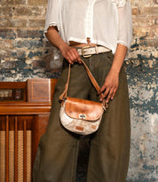 A person in a white blouse and olive pants holds the Bed|Stü Elect, a small brown and white shoulder bag, standing in front of a weathered brick wall and vintage wooden radio, capturing an effortless sense of vintage charm.