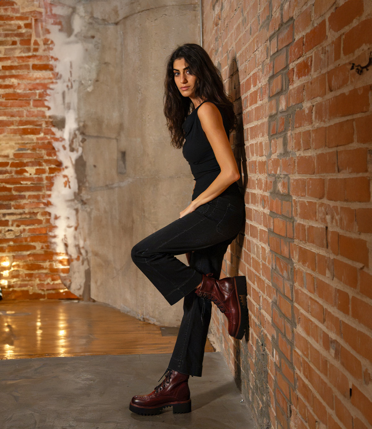 A woman in a black top and dark jeans leans against a brick wall, wearing dark red Bed|Stü Elisha Hi platform shoes with a lace-up profile and thick soles.