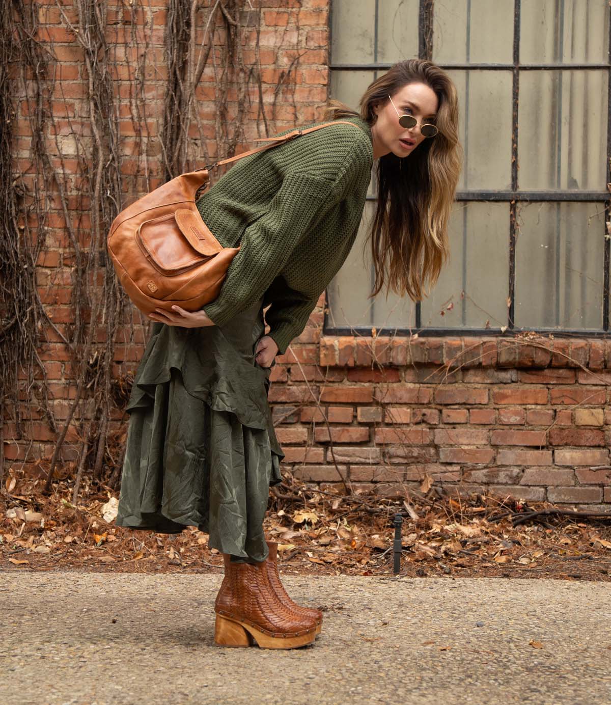 A woman in an olive green sweater and skirt, sunglasses, and a large brown shoulder bag stands outdoors by a brick wall, leaning forward in her Ellasphin sculptural style platform boots by Bed|Stü—her look elevated by the artisan fashion sensibility of this distinctive footwear.
