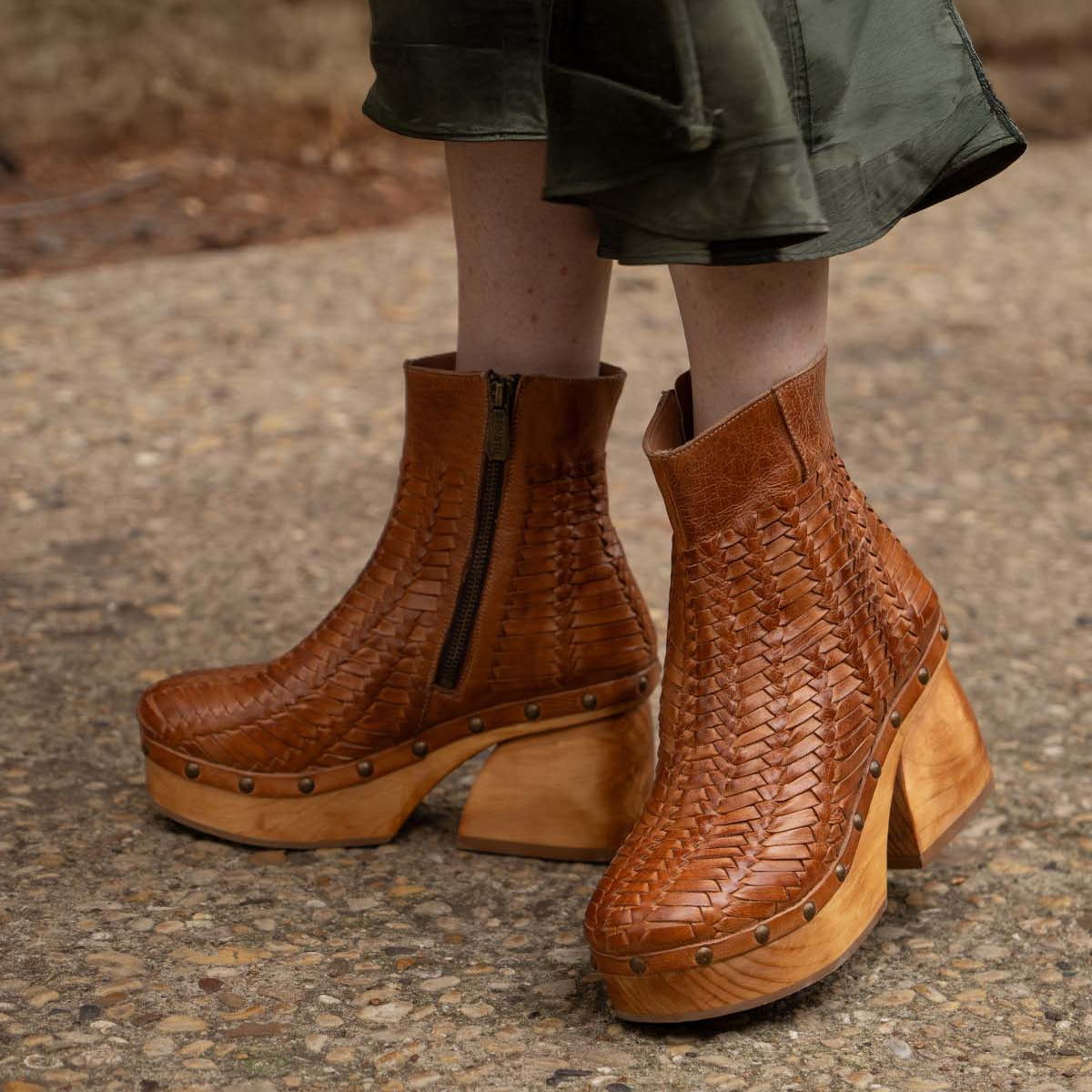 A person wearing the Ellasphin handwoven platform shoes by Bed|Stü, featuring brown leather, wooden soles, and brass studs, stands on a gravel path; only the lower legs and feet are shown, drawing attention to this distinctive piece of artisan footwear.