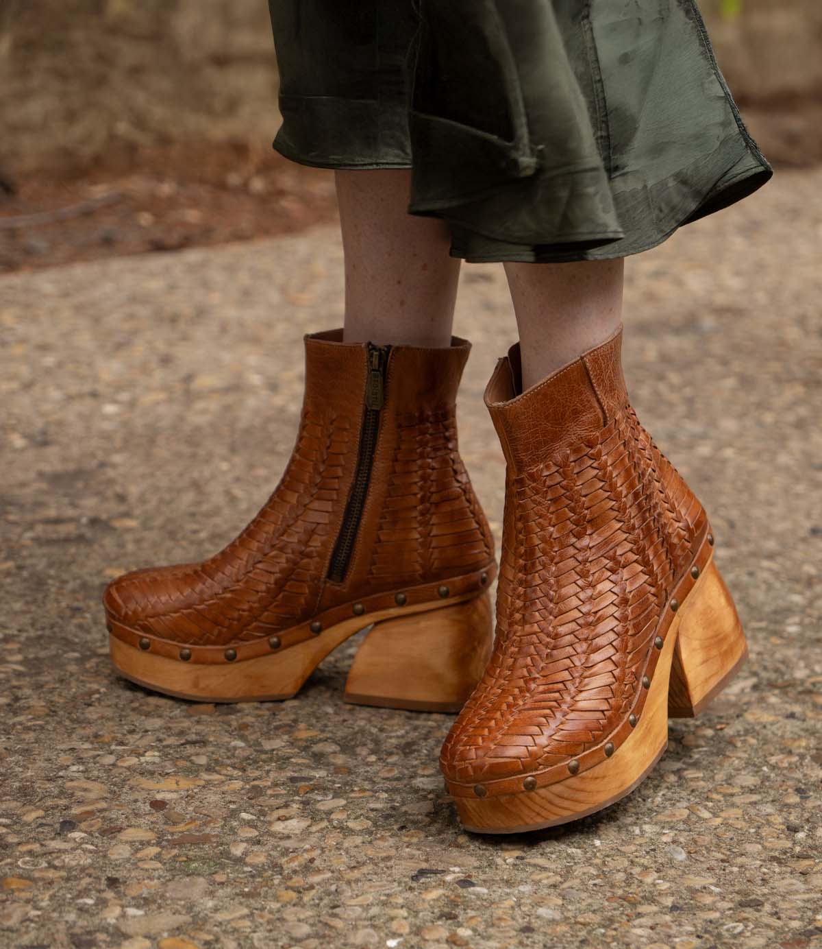 A person wearing the Ellasphin handwoven platform shoes by Bed|Stü, featuring brown leather, wooden soles, and brass studs, stands on a gravel path; only the lower legs and feet are shown, drawing attention to this distinctive piece of artisan footwear.