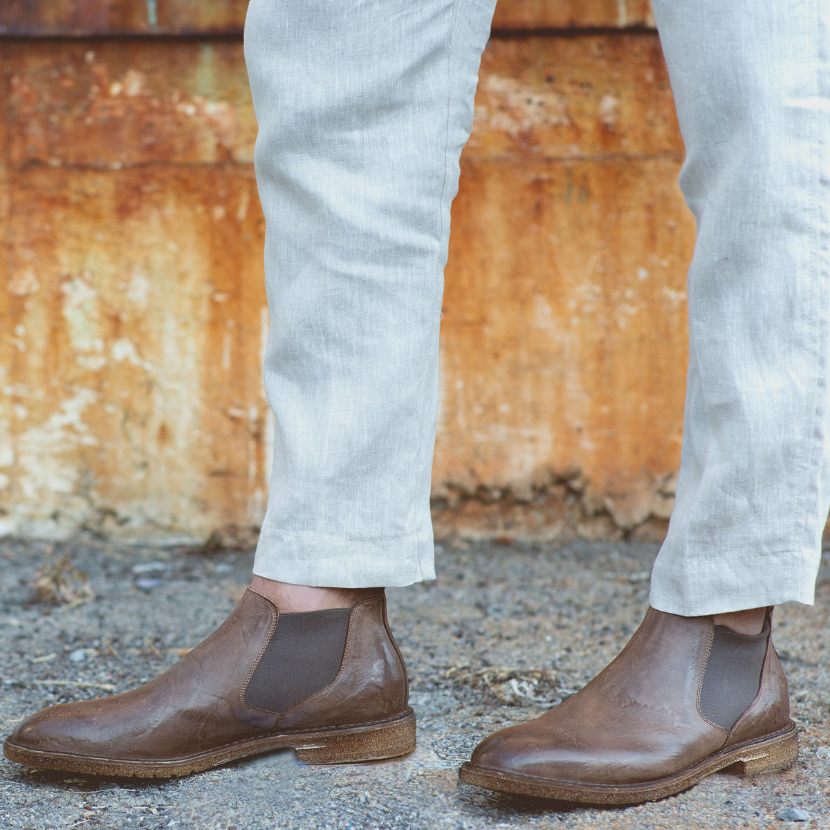 Wearing light-colored pants and Bed|Stü Elude Italian handcrafted brown boots, a person stands on gravel before a rusted metal surface.