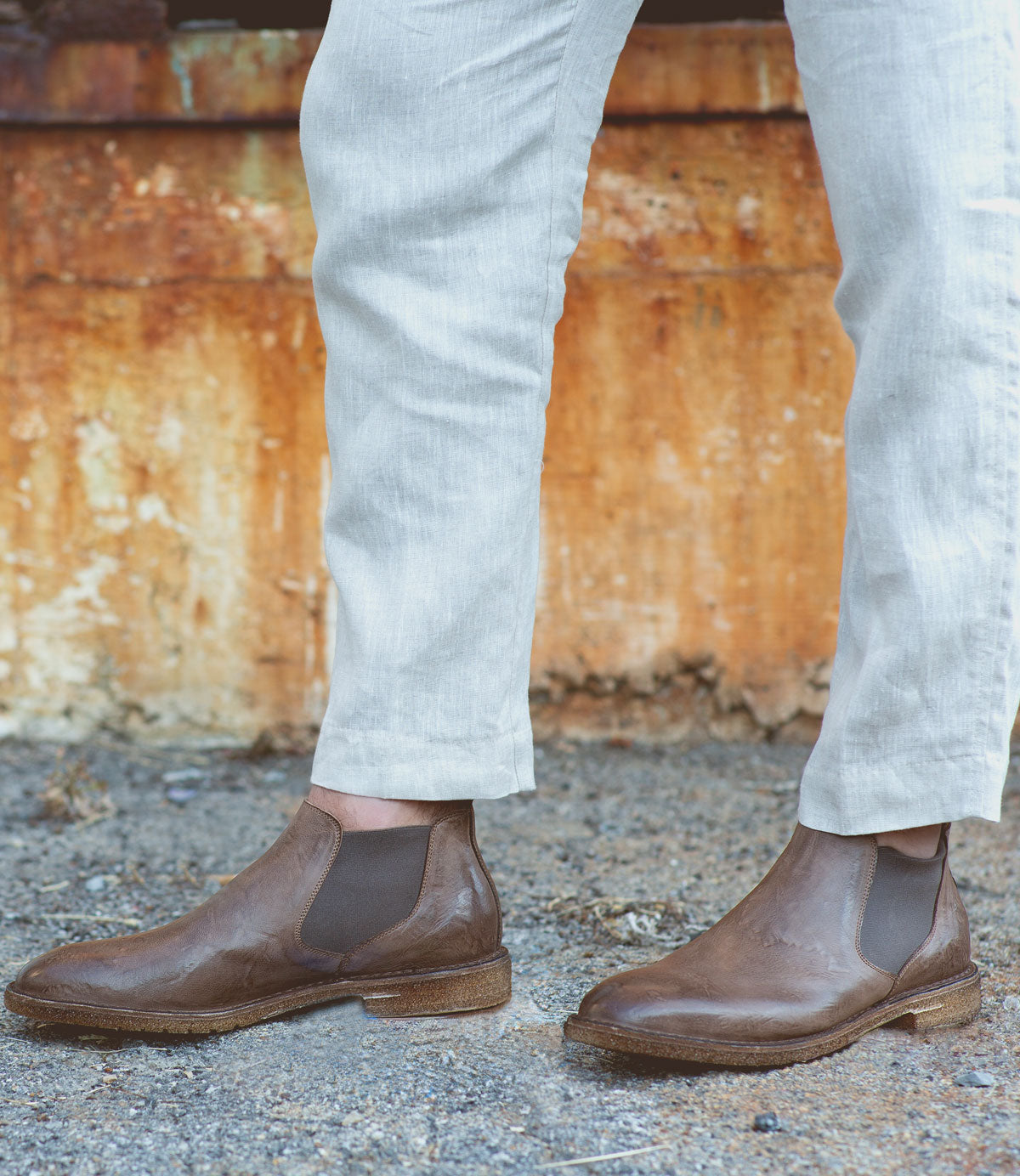 Wearing light-colored pants and Bed|Stü Elude Italian handcrafted brown boots, a person stands on gravel before a rusted metal surface.