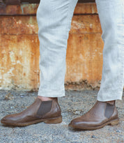 Wearing light-colored pants and Bed|Stü Elude Italian handcrafted brown boots, a person stands on gravel before a rusted metal surface.