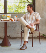 Wearing the stylish Elude men's leather boots by Bed|Stü, a man in a striped shirt and light pants sits by a table with oranges, drinks, and bread near a window in a rustic room.