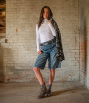 A woman stands in a rustic room wearing a white turtleneck, denim shorts, and the Bed|Stü Endure ankle strap boots featuring a durable treaded outsole, holding a patterned jacket over her shoulder.
