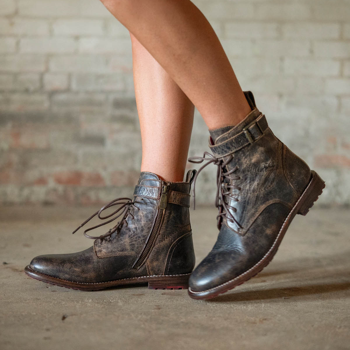 A person wearing the Bed|Stü Endure distressed brown rugged leather boots with buckles and a durable treaded outsole, standing on a concrete floor in front of a brick wall.