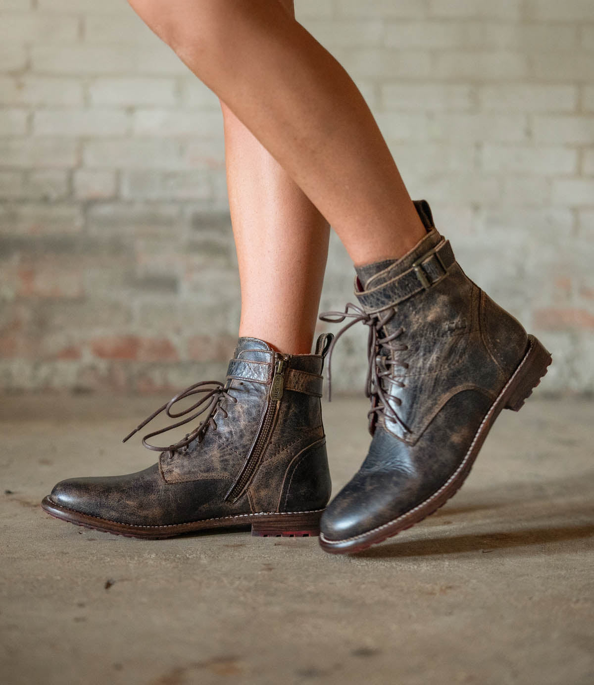 A person wearing the Bed|Stü Endure distressed brown rugged leather boots with buckles and a durable treaded outsole, standing on a concrete floor in front of a brick wall.