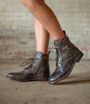 A person wearing the Bed|Stü Endure distressed brown rugged leather boots with buckles and a durable treaded outsole, standing on a concrete floor in front of a brick wall.