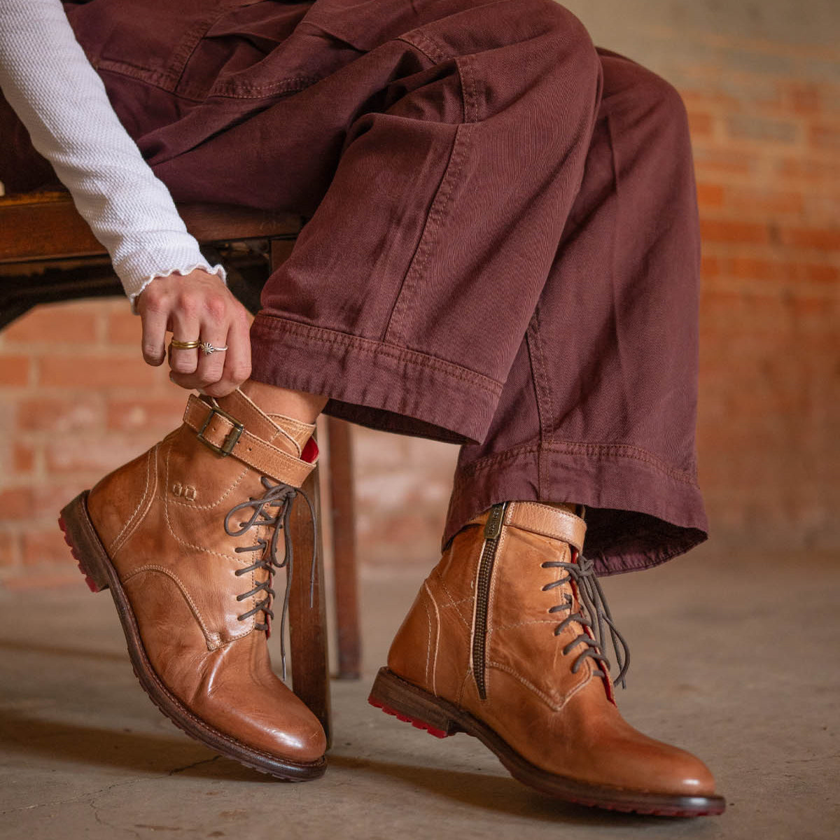 A person sits on a wooden chair, tying the laces of Bed|Stü’s Endure—a pair of rugged leather boots with a durable treaded outsole—while wearing maroon pants and a white ribbed long-sleeve shirt, in an indoor space with exposed brick walls.
