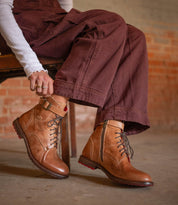 A person sits on a wooden chair, tying the laces of Bed|Stü’s Endure—a pair of rugged leather boots with a durable treaded outsole—while wearing maroon pants and a white ribbed long-sleeve shirt, in an indoor space with exposed brick walls.