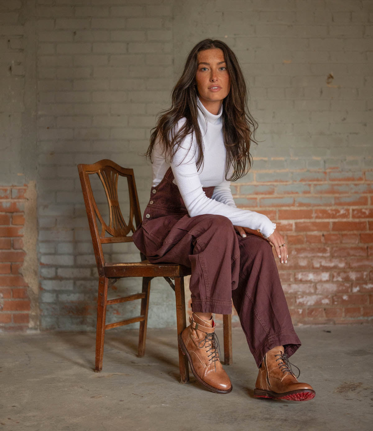 A woman with long brown hair sits sideways on a wooden chair, wearing a white turtleneck, brown overalls, and the Bed|Stü Endure rugged leather boots with a durable treaded outsole, in front of a brick and plaster wall.