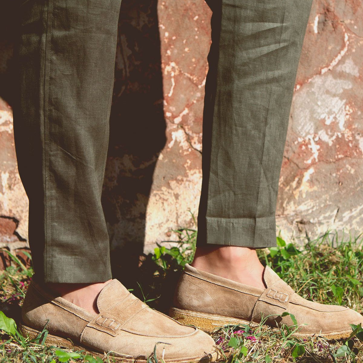 A person wearing Bed|Stü Envoy light brown handcrafted loafers and olive green pants stands on grass near a weathered stone wall.