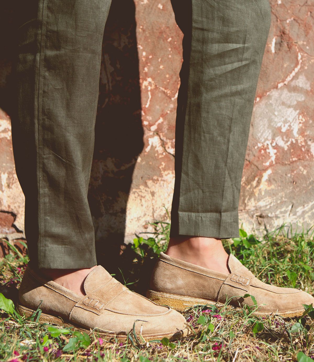 A person wearing Bed|Stü Envoy light brown handcrafted loafers and olive green pants stands on grass near a weathered stone wall.
