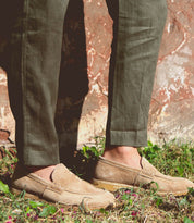 A person wearing Bed|Stü Envoy light brown handcrafted loafers and olive green pants stands on grass near a weathered stone wall.