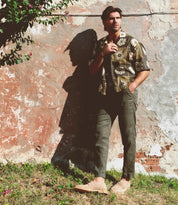 A man stands in sunlight against a weathered wall, wearing a patterned shirt, dark pants, and Bed|Stü's handcrafted Envoy loafers, one hand on his shirt collar and the other in his pocket.