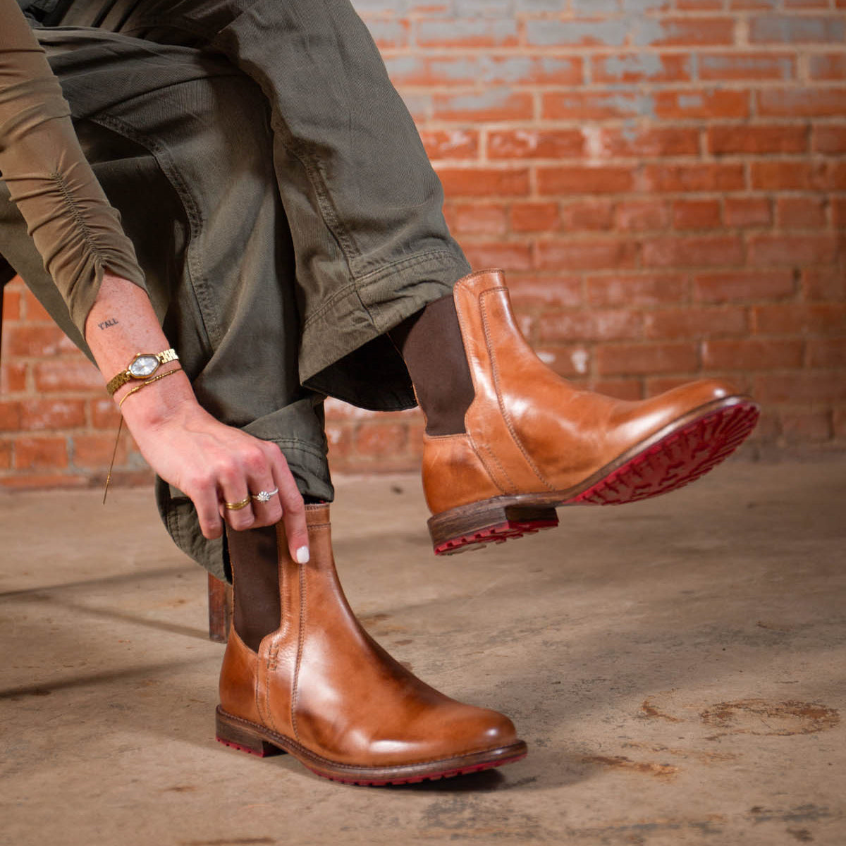 A person wearing the Ethic Chelsea boots by Bed|Stü—crafted from durable brown leather with distinctive red soles—paired with olive green pants, is seated on a chair against a brick wall background.
