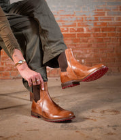 A person wearing the Ethic Chelsea boots by Bed|Stü—crafted from durable brown leather with distinctive red soles—paired with olive green pants, is seated on a chair against a brick wall background.