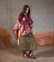A woman stands indoors in front of a painted brick wall, wearing a maroon patterned shawl, an olive green dress, and the Ethic durable, leather-lined brown Chelsea boots by Bed|Stü.