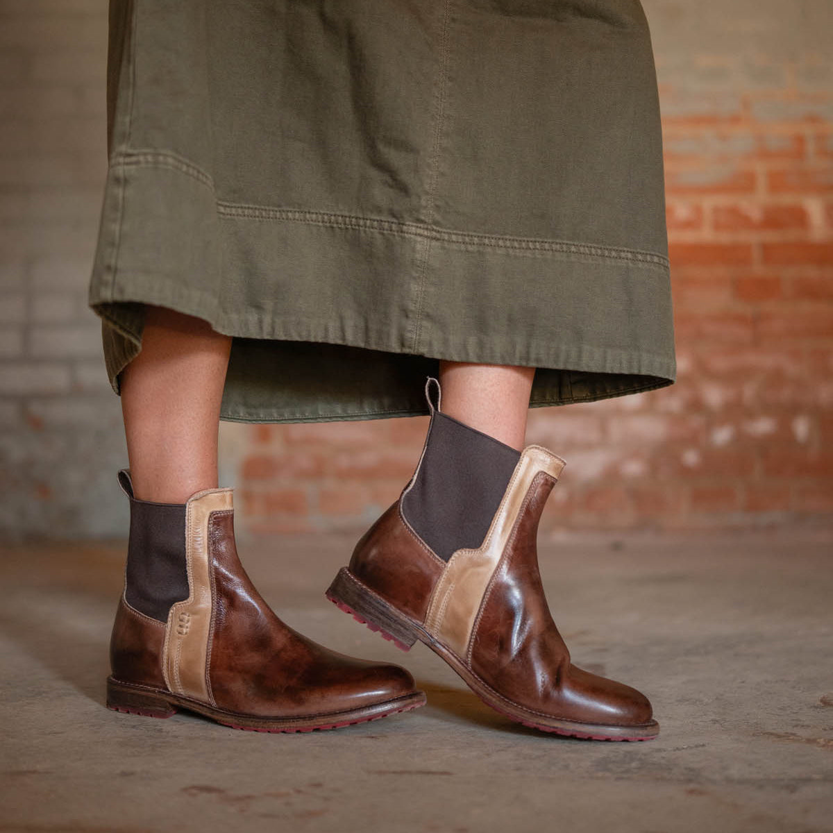 A person wearing the Bed|Stü Ethic durable brown leather Chelsea boots with black elastic panels and a long olive green skirt stands on a concrete floor, backed by a brick wall.