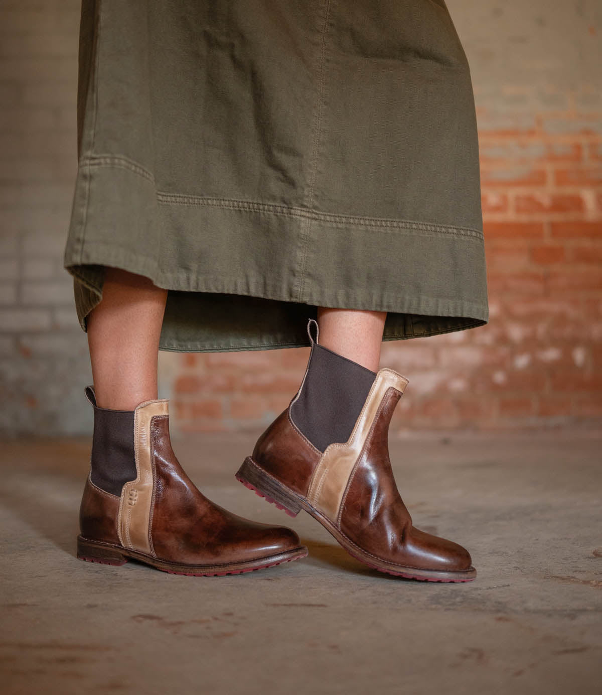 A person wearing the Bed|Stü Ethic durable brown leather Chelsea boots with black elastic panels and a long olive green skirt stands on a concrete floor, backed by a brick wall.