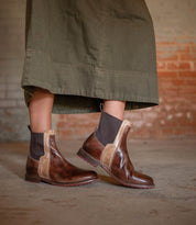 A person wearing the Bed|Stü Ethic durable brown leather Chelsea boots with black elastic panels and a long olive green skirt stands on a concrete floor, backed by a brick wall.