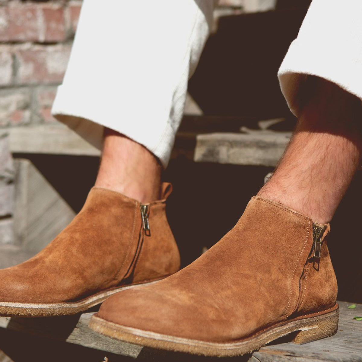 A person wearing Bed|Stü’s Evade men's suede boots with dual zippers sits on wooden steps, dressed in off-white pants. Handcrafted in Italy, the Evade boots add a refined touch to the casual scene.