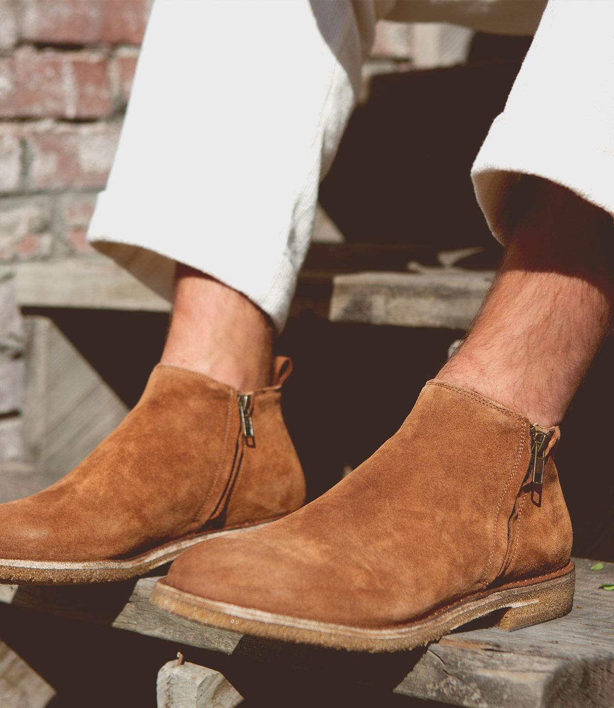 A person wearing Bed|Stü’s Evade men's suede boots with dual zippers sits on wooden steps, dressed in off-white pants. Handcrafted in Italy, the Evade boots add a refined touch to the casual scene.