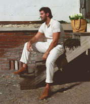 A man dressed in a cream outfit and wearing Bed|Stü’s Evade brown dual zipper boots, handcrafted in Italy, sits on wooden stairs outside, with a woven bag of flowers nearby.