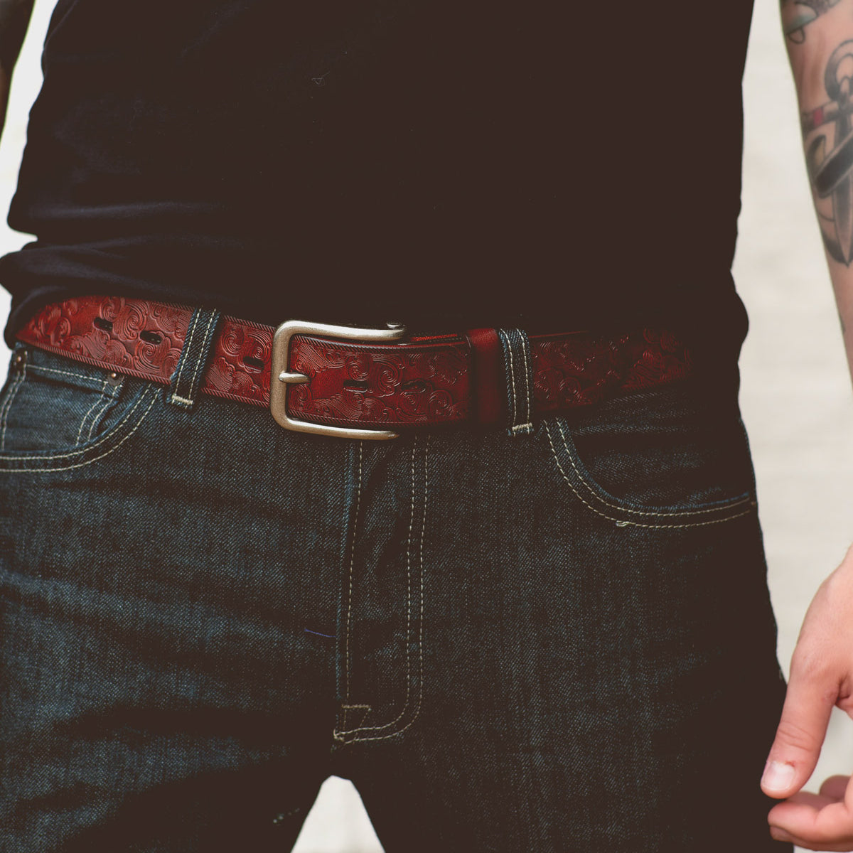 A person is wearing a black t-shirt, dark blue jeans with visible stitching, and the Bed|Stü Everton—a dark brown leather belt showcasing an intricate scroll pattern and finished with a silver removable buckle. One tattooed arm is partially visible.
