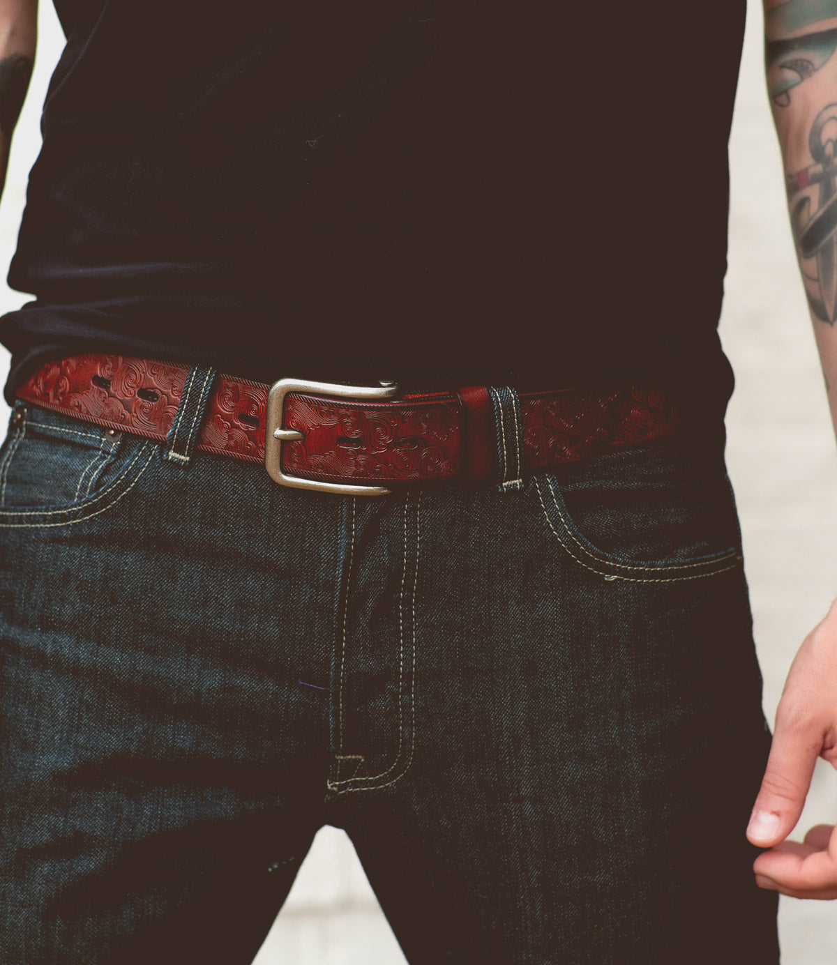 A person is wearing a black t-shirt, dark blue jeans with visible stitching, and the Bed|Stü Everton—a dark brown leather belt showcasing an intricate scroll pattern and finished with a silver removable buckle. One tattooed arm is partially visible.