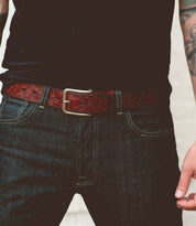 A person is wearing a black t-shirt, dark blue jeans with visible stitching, and the Bed|Stü Everton—a dark brown leather belt showcasing an intricate scroll pattern and finished with a silver removable buckle. One tattooed arm is partially visible.