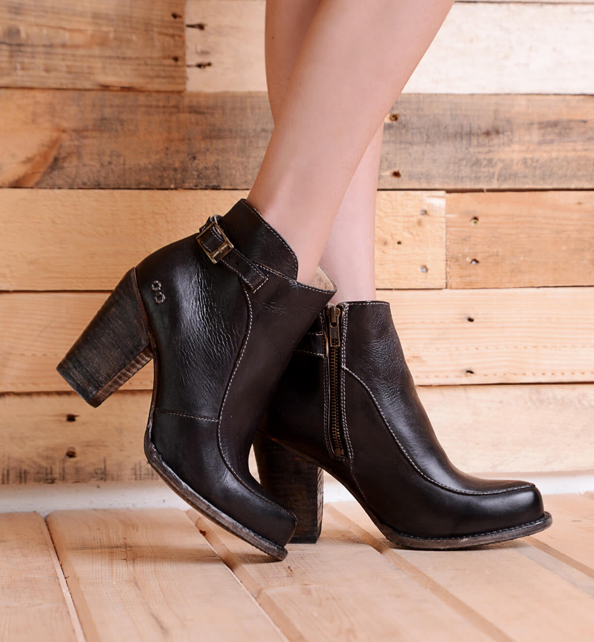 A person wearing the Isla black smooth leather ankle boots by Bed|Stü, featuring chunky heels, buckles, zippers, and a subtle ankle strap detail, stands on a wooden floor with a wooden plank wall in the background.