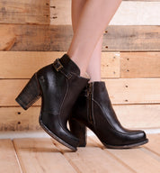 A person wearing the Isla black smooth leather ankle boots by Bed|Stü, featuring chunky heels, buckles, zippers, and a subtle ankle strap detail, stands on a wooden floor with a wooden plank wall in the background.